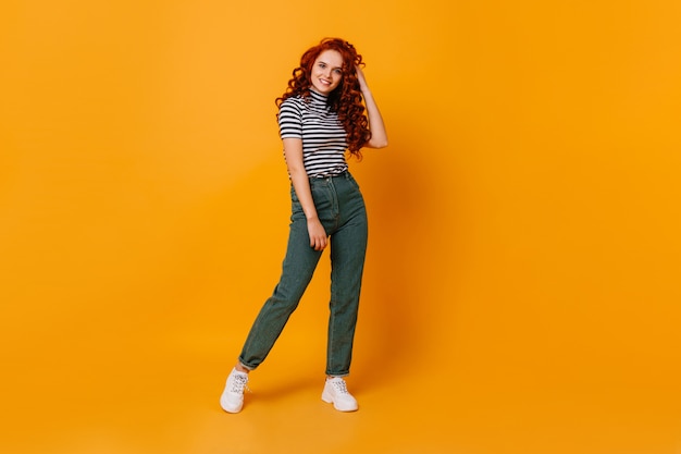 Bastante joven toca su pelo rojo rizado y sonríe. Retrato de niña en jeans y top en estudio naranja.