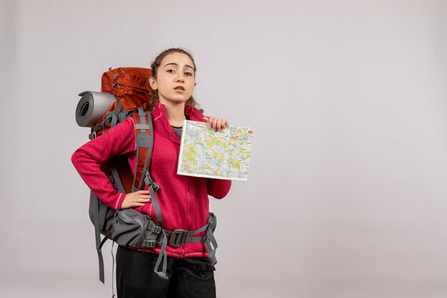 Bastante joven viajero con mochila grande sosteniendo el mapa poniendo la mano en la cintura en gris