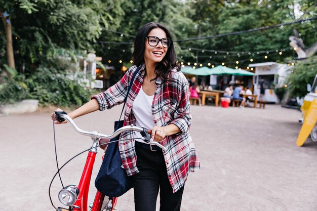 Bastante jovencita con cabello oscuro de pie en la calle con bicicleta. Foto de chica morena interesada en pantalones negros divirtiéndose en fin de semana.