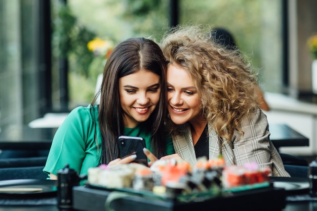 Bastante jovencitas, socios mirando el teléfono celular con plato de sushi en la mesa. Terraza del restaurante moderno.