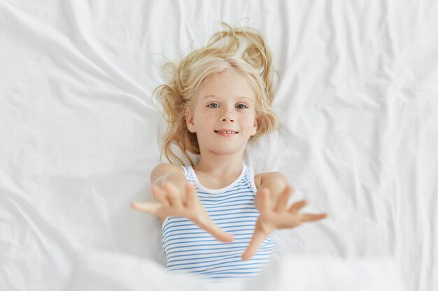 Bastante pequeño niño estirando sus manos mientras está acostado en la cama. Niña de ojos azules descansando en la cama
