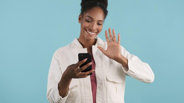 Bastante sonriente chica afroamericana casual saludando felizmente la mano hola en el teléfono inteligente sobre fondo colorido