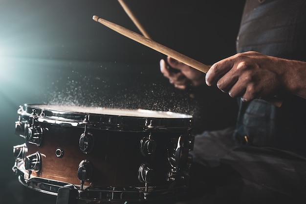 El baterista con baquetas golpeando el tambor con salpicaduras de agua sobre fondo negro bajo iluminación de estudio de cerca.