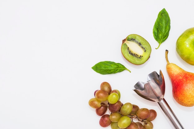 Una batidora eléctrica con kiwi; manzana; Pera; Uvas y hojas de albahaca sobre fondo blanco