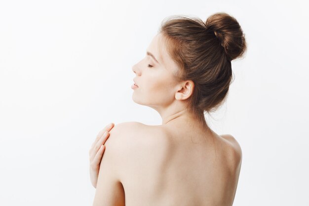 Bauty y salud. Guapa, delgada, sana y caucásica, con cabello oscuro en peinado de moño y piel desnuda, se da vuelta con la espalda, toca los hombros con las manos, tiene una mirada relajada y sutis