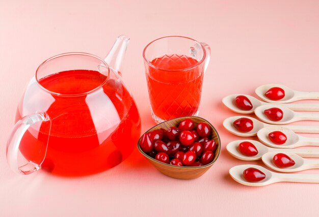 Bayas de cornejo en cucharas de madera y cuenco con bebidas vista de ángulo alto en rosa