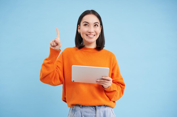 Photo Gratis beautiful young korean woman with tablet pointing finger up and looking on top advertisement showing