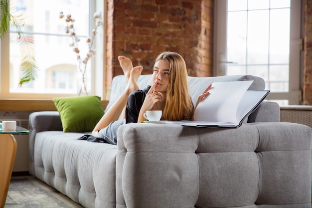 Beauty Day para ti. Mujer vestida con bata de seda haciendo su rutina diaria de cuidado de la piel en casa. Tumbado en el sofá, leyendo la revista, tomando café, pensativo. Concepto de belleza, cuidado personal, cosmética, juventud.