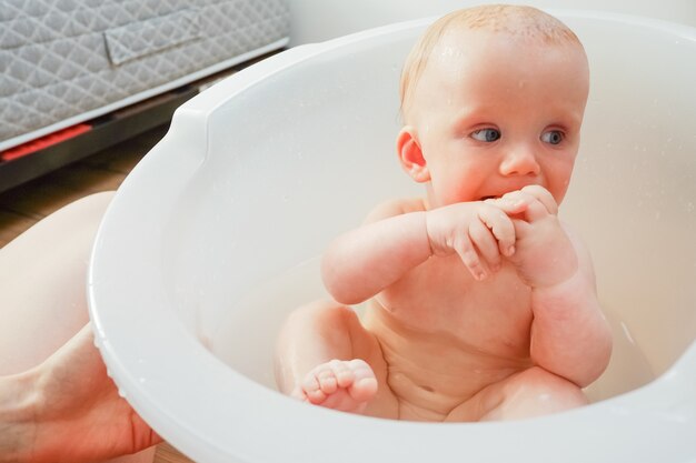 Bebé adorable pensativo que muerde el juguete de goma mientras tiene bañera en casa. Madre bañando al niño en la bañera en el interior de la casa. Fotografía de cerca. Concepto de cuidado infantil o salud