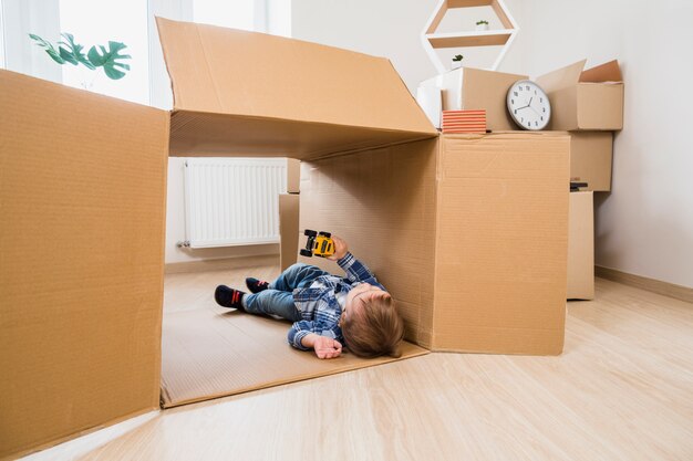 Bebé adorable que miente en la caja de cartón que juega con el coche del juguete en casa