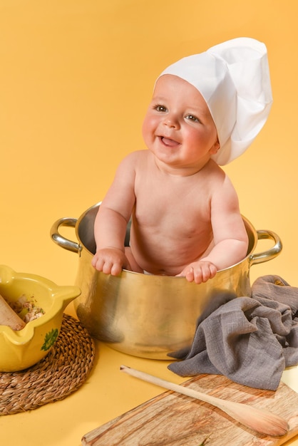 Bebé caucásico sonriente escondido dentro de una olla con gorro de cocinero y utensilios de cocina sobre fondo amarillo