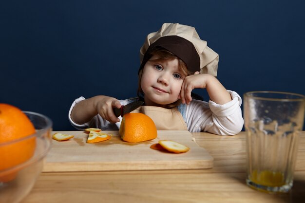 Bebé chef en elegante delantal y sombrero de pie en la mesa con tabla de cortar de madera, usando un cuchillo afilado mientras corta naranjas frescas para ensalada. Imagen de adorable niña ayudando a la madre en la cocina