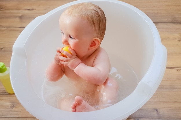 Bebé mojado lindo pensativo que muerde el juguete de goma amarillo mientras está sentado y se baña en la bañera en casa. Fotografía de cerca. Concepto de cuidado infantil o salud