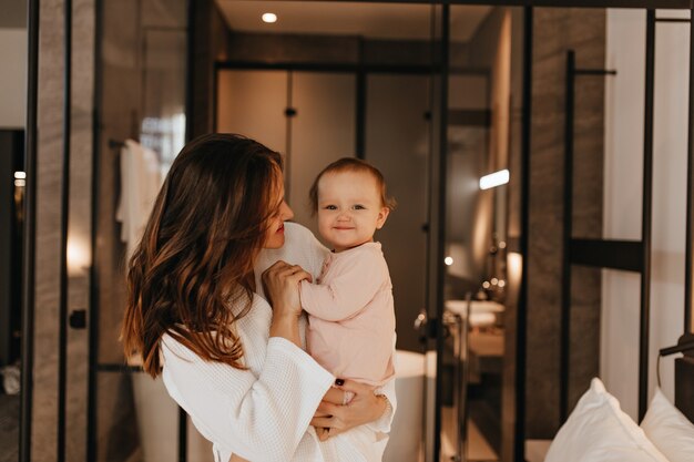 Bebé en mono rosa sonríe dulcemente mientras su mamá le habla. Señora de pelo largo en bata de baño blanca jugando con el niño en el fondo del baño.