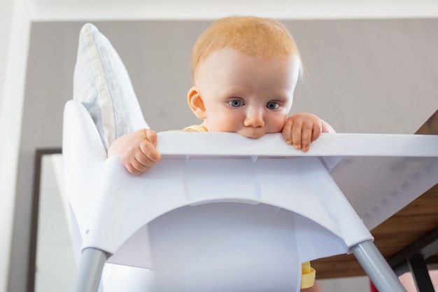 Foto gratuita bebé pelirrojo pensativo mirando hacia abajo en el suelo desde la silla alta y la bandeja de morder. ángulo bajo. proceso de alimentación o concepto de cuidado infantil