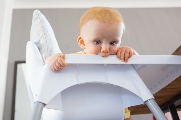 Bebé pelirrojo pensativo mirando hacia abajo en el suelo desde la silla alta y la bandeja de morder. Ángulo bajo. Proceso de alimentación o concepto de cuidado infantil