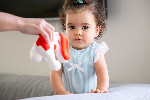 Bebé de pelo rizado oscuro pensativo mientras su mamá le da un juguete rojo y blanco al niño. Niño en casa y concepto de infancia.