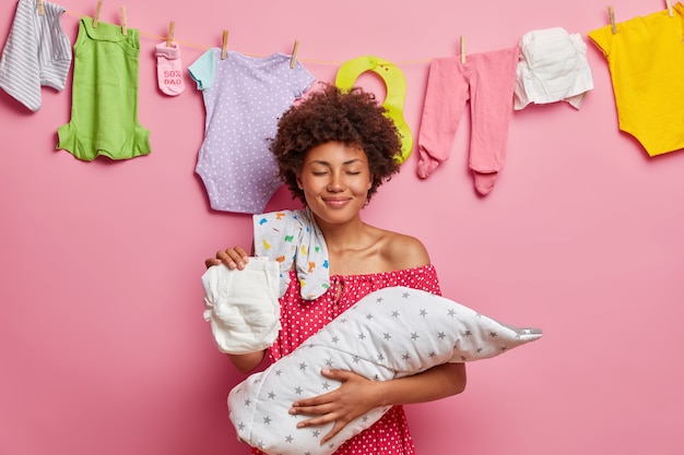 Bebé recién nacido descansa en manos de las madres. La madre cariñosa de la mujer de pelo rizado complacida sostiene al bebé que duerme envuelto en una manta en las manos tiene un mono del pañal en poses del hombro interior. Concepto de familia feliz.