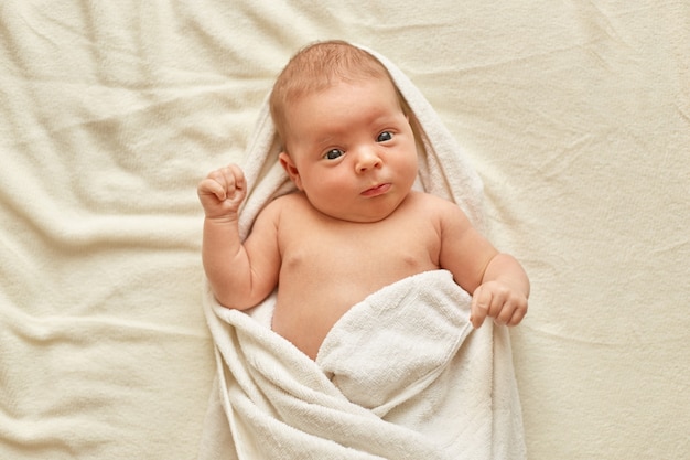 Bebé recién nacido con toalla después del baño, lindo niño pequeño acostado en la cama sobre una manta blanca, mirando y estudiando cosas externas. Cuidado de la infancia y el bebé.