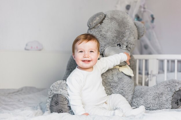 Bebé rubio adorable sobre cama blanca con oso de peluche