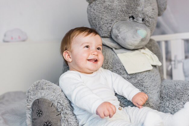 Bebé rubio adorable sobre cama blanca con oso de peluche
