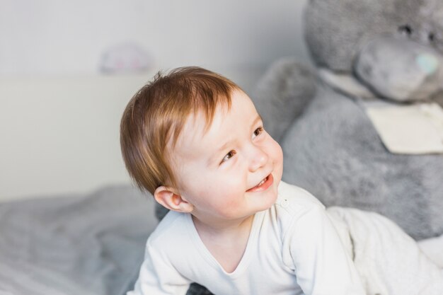 Bebé rubio adorable sobre cama blanca con oso de peluche