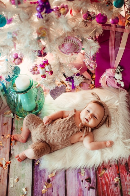 Bebé en traje de ciervo se encuentra en la almohada blanca suave bajo el árbol de Navidad blanco con juguetes de color rosa