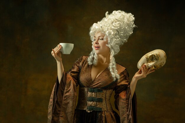 Beber café con mascarilla. Retrato de mujer joven medieval en ropa vintage marrón en la pared oscura. Modelo femenino como duquesa, persona real. Concepto de comparación de épocas, moderno, moda.