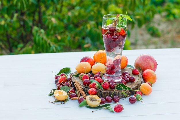 Beber en un vaso con frutas, especias, vista lateral de la tabla de cortar sobre fondo de madera y jardín