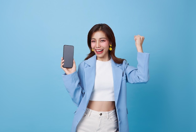 Bella adolescente asiática sosteniendo una maqueta de teléfono inteligente de pantalla en blanco y celebrando con la mano en alto