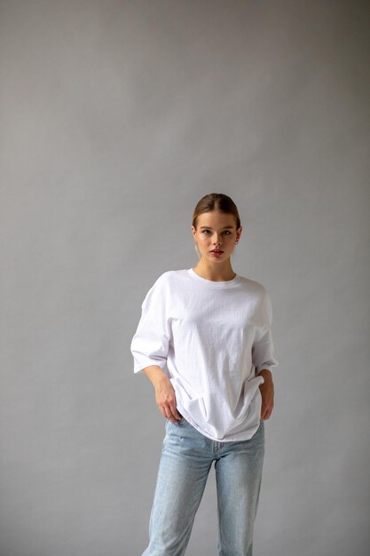 Bella chica rubia con una camiseta blanca de gran tamaño y vaqueros azules posando sobre un fondo gris