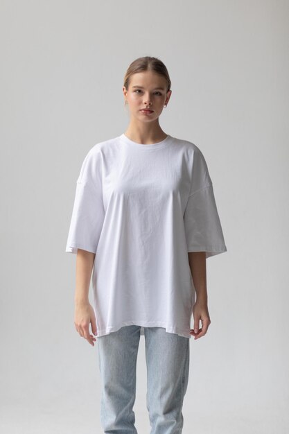 Bella chica rubia con una camiseta blanca de gran tamaño y vaqueros azules posando sobre un fondo gris