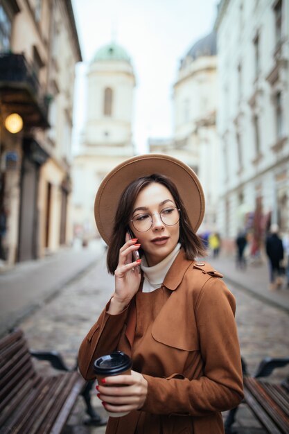 Bella dama con abrigo marrón hablando por teléfono móvil caminando al aire libre en un frío día de otoño