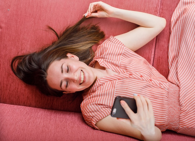 Bella dama acostada, mirando su teléfono y sonriendo en la habitación en un sofá con vestido rosa a rayas