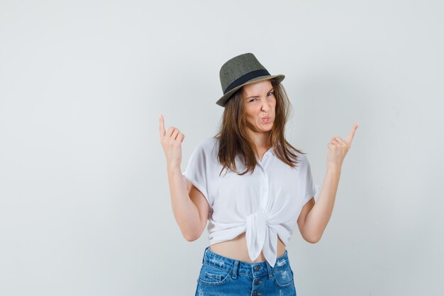 Bella dama apuntando hacia arriba mientras hace pucheros con sus labios en blusa blanca, sombrero, vista frontal.