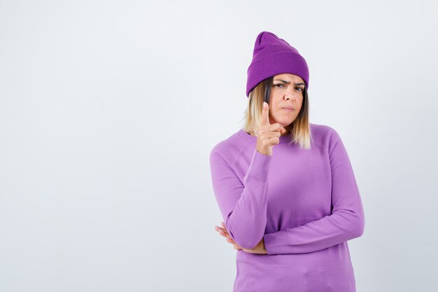 Bella dama apuntando hacia arriba con suéter, gorro y mirando confiada, vista frontal.