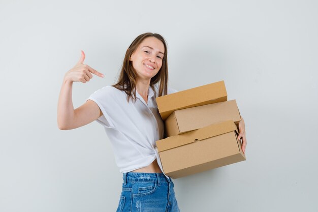 Bella dama apuntando a cajas en blusa blanca y mirando complacida. vista frontal.