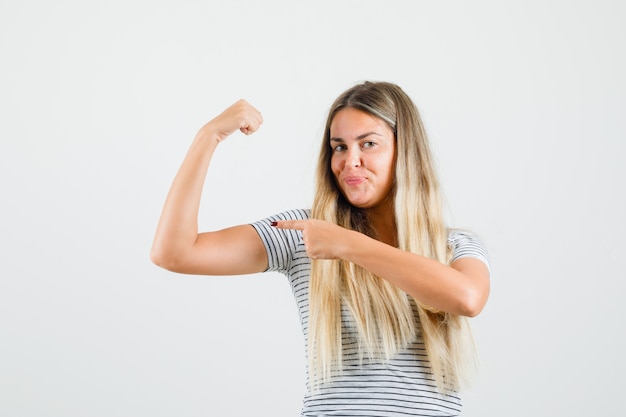 Foto gratuita bella dama apuntando a los músculos de su brazo en camiseta y mirando confiada, vista frontal.