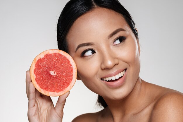 bella dama asiática mirando a un lado y sosteniendo una rodaja de pomelo cerca de la cara