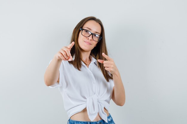 Bella dama en blusa blanca, gafas apuntando y mirando seguro, vista frontal.