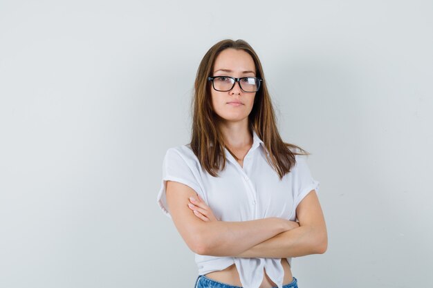 Bella dama en blusa blanca, gafas de pie con los brazos cruzados y mirando atenta, vista frontal.