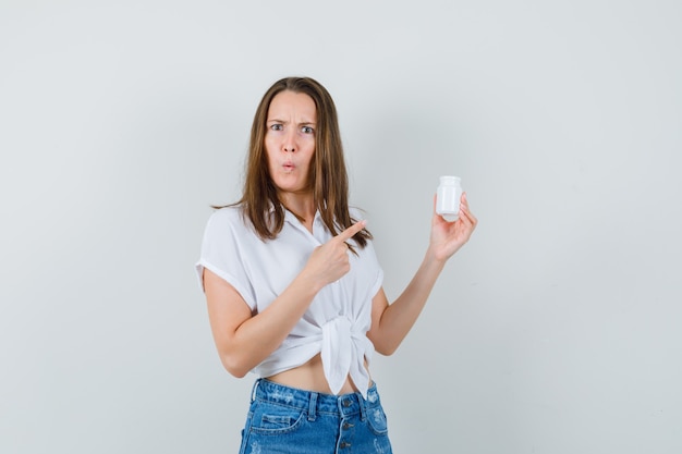 Bella dama en blusa blanca, jeans mirando una botella de píldoras y mirando confundida, vista frontal.