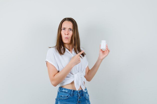 Bella dama en blusa blanca, jeans mirando una botella de píldoras y mirando confundida, vista frontal.