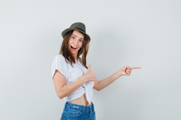 Bella dama en blusa blanca, sombrero apuntando a un lado mientras muestra el pulgar hacia arriba y mira alegre, vista frontal.