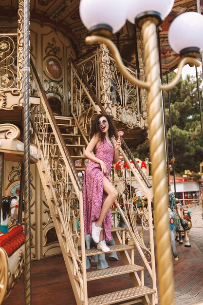 Bella dama con cabello oscuro y rizado con gafas de sol y vestido de pie con caramelos en la mano y felizmente mirando a un lado mientras pasa tiempo en un hermoso carrusel en el parque de diversiones