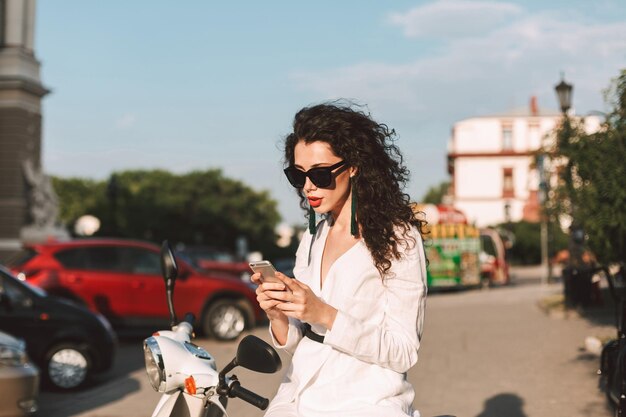 Bella dama con cabello oscuro y rizado en traje blanco y gafas de sol sentada en un ciclomotor blanco y cuidadosamente usando su teléfono celular mientras pasa tiempo en la calle de la ciudad