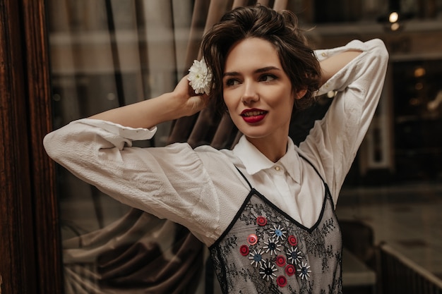 Bella dama con cabello rizado y labios rojos mirando hacia adentro. Mujer morena fresca en blusa ligera con flores posando en el café.