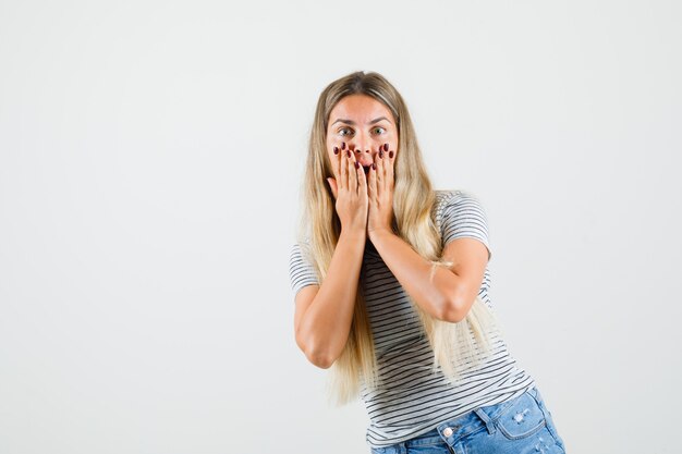 Bella dama en camiseta sosteniendo la mano en las mejillas y mirando asustada, vista frontal.