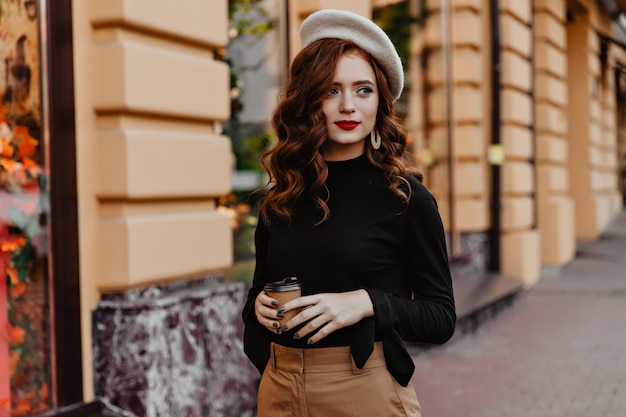 Bella dama francesa con taza de café mirando a su alrededor. Chica rizada pensativa en blusa negra caminando por la calle.