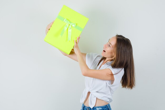 Bella dama levantando caja de regalo en blusa blanca, pantalón de jeans, vista frontal.